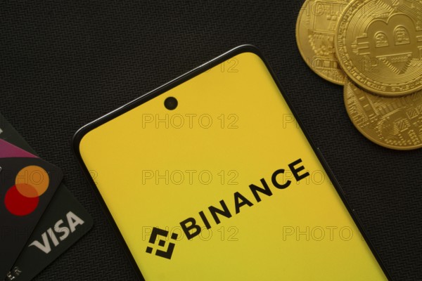 Dhaka, Bangladesh- 22 June 2025: Smartphone displaying Binance logo surrounded by Bitcoin coins and payment cards