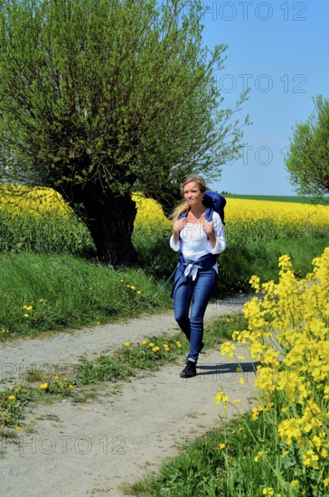 Young woman with backpack walking on a countryroad with willow trees and field of rape in Skivarp, Skåne County, South Sweden