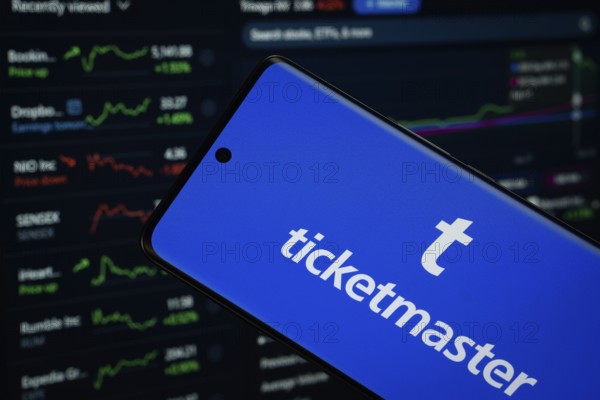 Dhaka, Bangladesh- 18 March 2025: Ticketmaster logo is displayed on a smartphone screen, with a stock graph webpage visible on a computer screen in the background