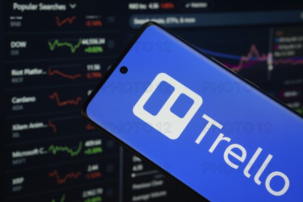 Dhaka, Bangladesh- 18 March 2025: trello logo is displayed on a smartphone screen, with a stock graph webpage visible on a computer screen in the background