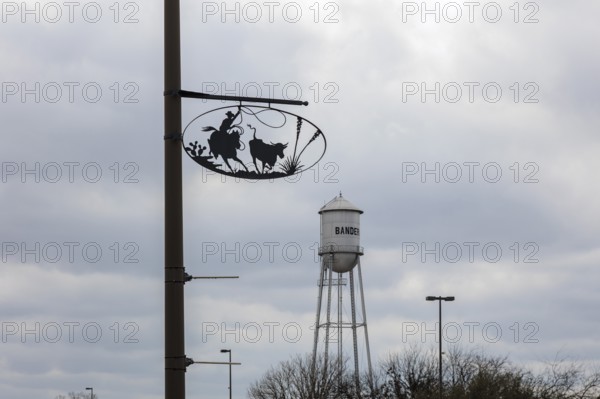 Bandera water tower behind artistic metal sign showing cowboy life on pole in Bandera, Texas, USA