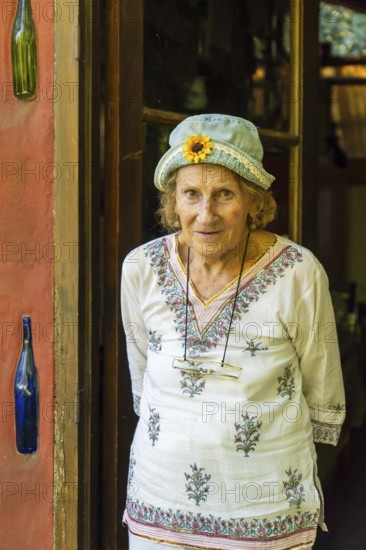Elderly woman wearing a denim hat with a sunflower decoration and a white tunic smiles warmly at the doorway of her uniquely decorated home, exuding a sense of contentment and peace