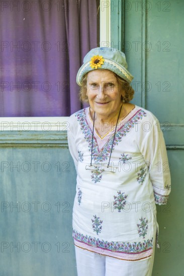 Portrait of a cheerful senior woman wearing a denim hat with a decorative sunflower, standing in front of a turquoise wall with a window and purple curtain