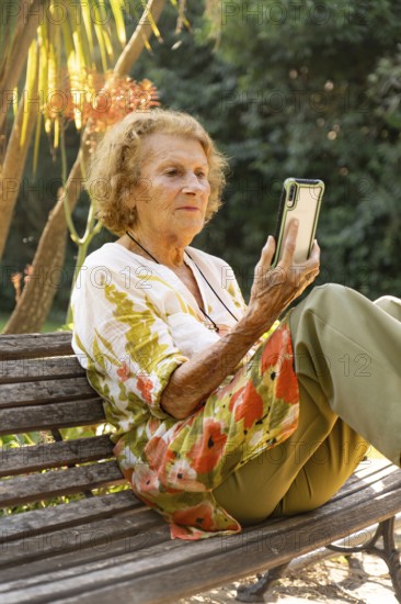 Elderly woman enjoying a tranquil moment in the park, sitting comfortably on a bench while browsing her smartphone and connecting with the world around her under the warm sunlight