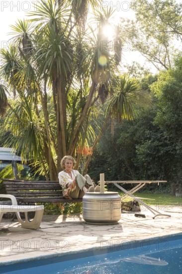 Elderly woman enjoys a peaceful moment in her backyard, sitting on a bench next to a sparkling pool, surrounded by lush greenery and bathed in the warm afternoon sun