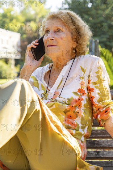Senior woman sitting comfortably on a park bench, making a phone call and enjoying the warm summer weather while staying connected with loved ones in a serene outdoor setting