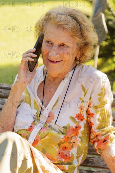 Elderly woman with glasses hanging from a string around her neck is sitting on a wooden bench outside in a garden, smiling while talking on her smartphone