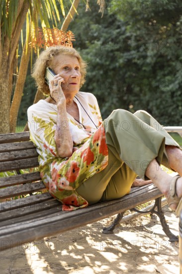Senior woman sitting comfortably on a park bench, enjoying a sunny day while making a phone call. Embracing the outdoors, she stays connected and relaxed in nature's tranquility