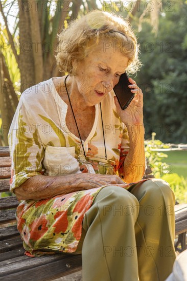 Senior woman sitting on a wooden bench in a serene garden, using her smartphone to enjoy communication and stay connected during her retirement, embracing a peaceful lifestyle in nature