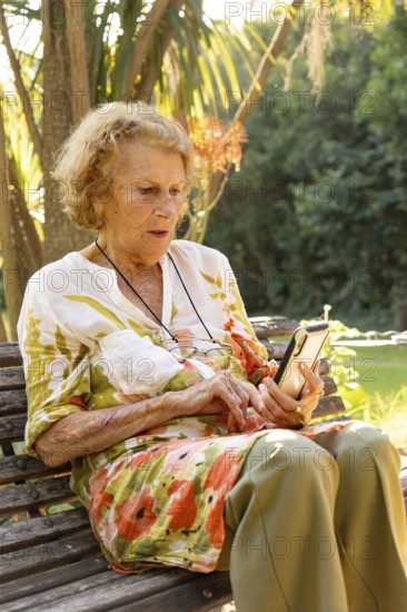 Senior woman sitting on a wooden bench in a park, enjoying the pleasant weather while using her smartphone, showcasing the intersection of technology and aging