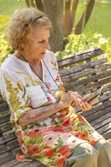 Senior woman sitting on a wooden bench in a park, enjoying the warmth of summer while using her smartphone to stay connected with family and friends in the digital age