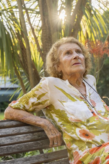 Senior woman enjoying a tranquil moment in a lush garden, sitting on a wooden bench while sunlight gently filtering through vibrant leaves, embracing the peace of nature