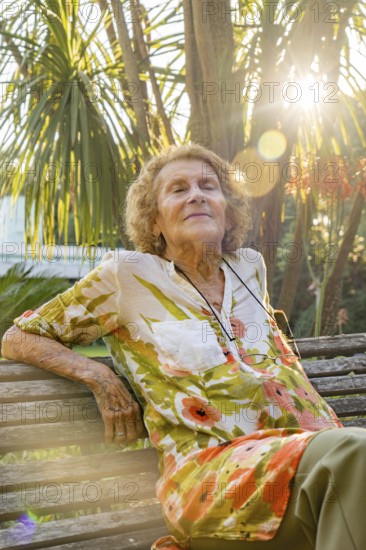 Elderly woman with closed eyes enjoying sunlight while sitting on wooden bench in lush botanical garden, embracing tranquility and nature's beauty