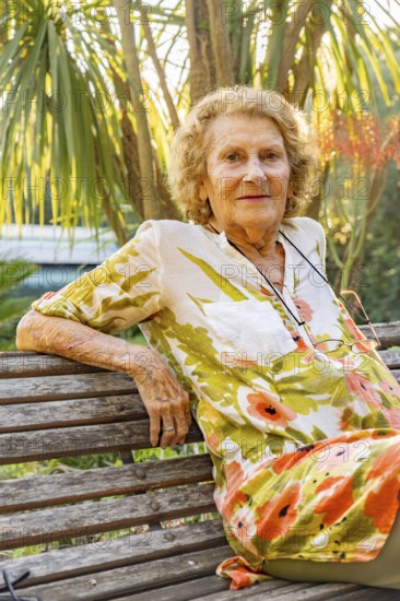 Smiling senior woman relaxing on a wooden bench amidst a vibrant tropical garden, soaking in the peaceful atmosphere and warm sunlight, embodying the essence of leisure and contentment