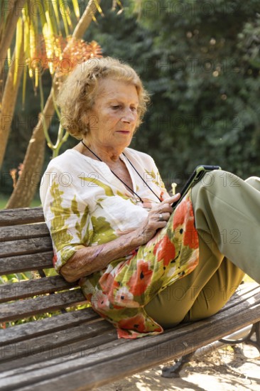 Senior woman sitting comfortably on a wooden bench in a park, using a tablet while enjoying the serene surroundings filled with greenery and sunlight, embracing a moment of relaxation