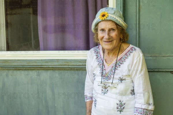 Elderly woman smiling while leaning against a light green wooden door, wearing a light blue hat with a sunflower and a white floral-patterned shirt by a window with purple curtains