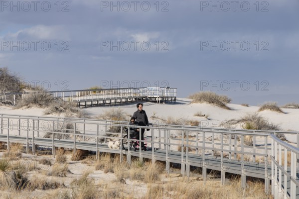 Man and handicapped woman in wheelchair stroll down the Interdune Boardwalk with their ped dog at White Sands Nationl Park near Alamogordo, New Mexico