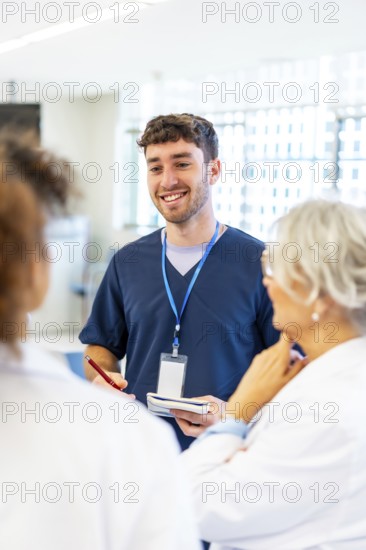 Young male nurse smiling and taking notes while talking with medical team in a hospital hallway