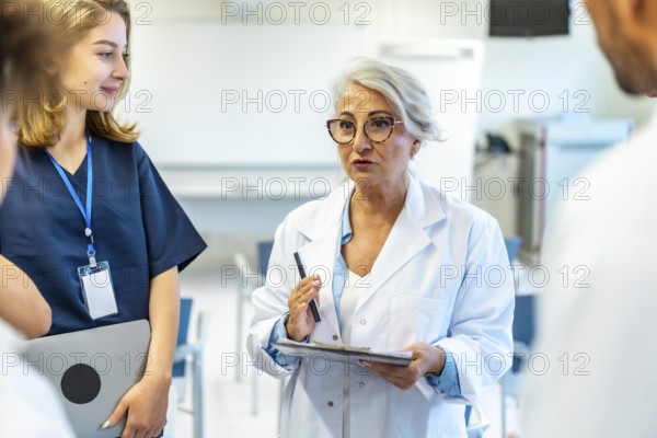 Experienced female doctor leading a medical briefing, holding a clipboard and pen, addressing colleagues in a hospital conference room