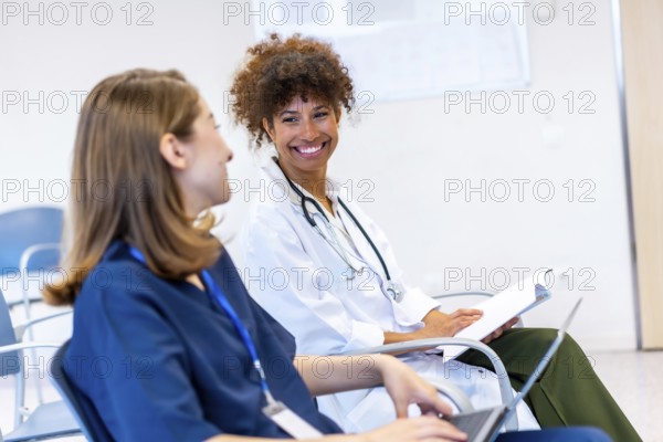 Two female doctors are sitting and smiling while discussing medical information during a conference in a hospital waiting room