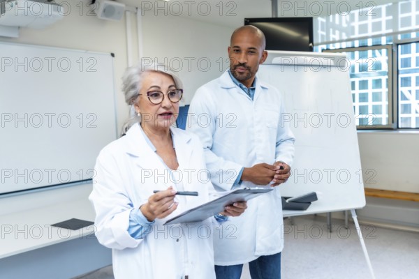 Senior female doctor explaining a healthcare project to colleagues, holding clipboard and pointing with a pen, standing next to a male doctor in a conference room