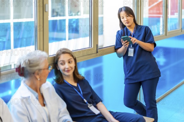 Asian nurse using a smartphone while colleagues chat and relax during a break in the hospital corridor, enjoying moments of teamwork