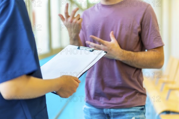 Young man explaining his symptoms using gestures to a nurse holding a medical chart in a hospital corridor