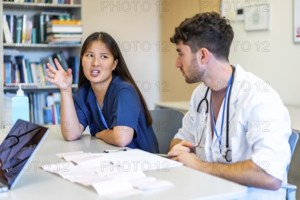 Two young doctors reviewing medical records and having a discussion about a patient chart at a library table in a hospital