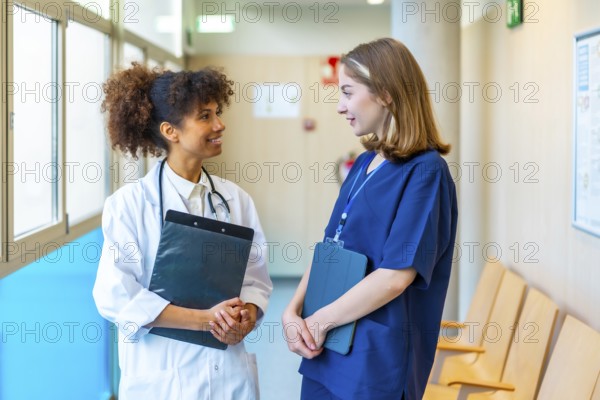 Female doctor and nurse engaging in a friendly conversation while reviewing medical records in a bustling hospital corridor
