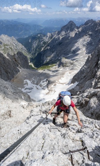 Mountaineer with helmet climbing in a secured via ferrata, Zugspitz via ferrata, ascent to the Zugspitze, view of mountain basin with snowfield of the Höllentalferner and Höllental, Wetterstein range, Wetterstein range, Bavaria, Germany
