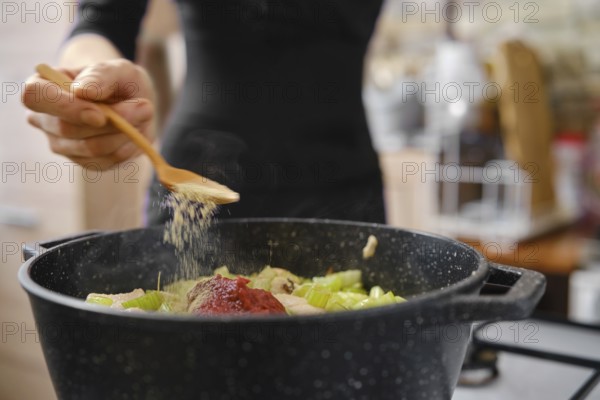 Unrecognizable woman sprinkling spice over vegetables in a black pot while preparing a hearty meal