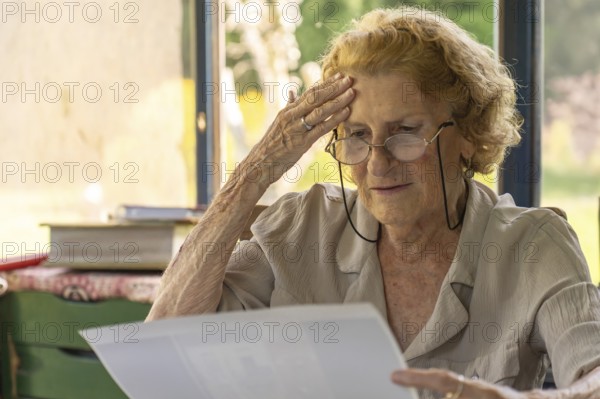 Senior woman wearing glasses, reading a letter at home and feeling a wave of worry, touching her forehead with one hand, displaying a mix of concern and confusion