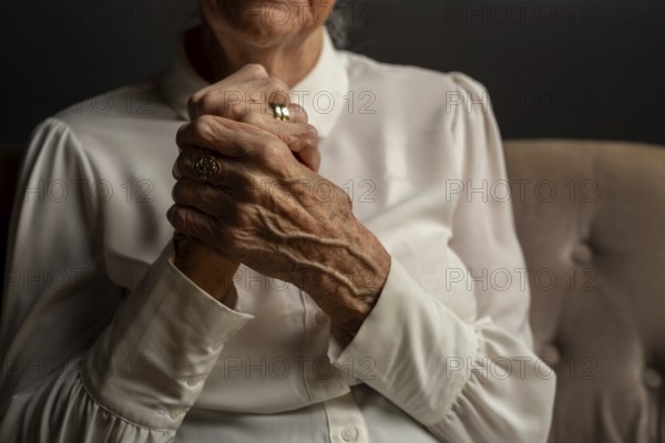 90 year old woman in white blouse holding folded hands, conveying thoughtfulness in a dim light, old age, symbolic image, Germany