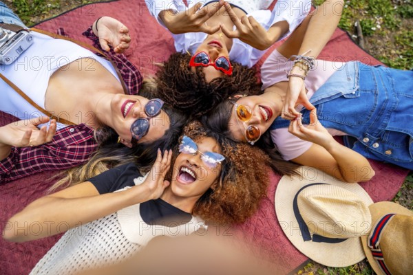 Four multi ethnic female friends are lying in circle on blanket in a park, making heart shape with hands and taking selfie