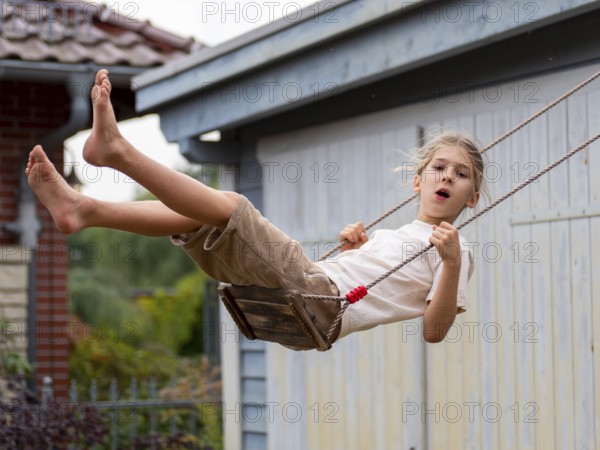Boy swinging while sitting on children's swing at shed and garden gate, Germany
