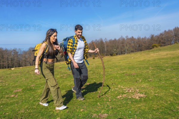 Young couple walking hand in hand across a vibrant green meadow, carrying backpacks and using a hiking stick under a sunny sky