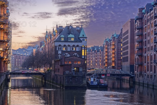 The moated castle in Hamburg's Speicherstadt between the Holländischbrookfleet and the Wandrahmfleet with the bridges of the Servant Row. At dusk, windows are illuminated. Speicherstadt, Hamburg, Germany