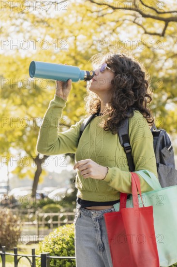 Woman with curly hair and sunglasses drinks from a blue water bottle in a park. She carries reusable bags and wears a green sweater, embodying an eco-friendly lifestyle on a sunny day