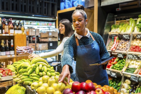 Supermarket employee using digital tablet while arranging fresh fruit on display, customer choosing bananas in background