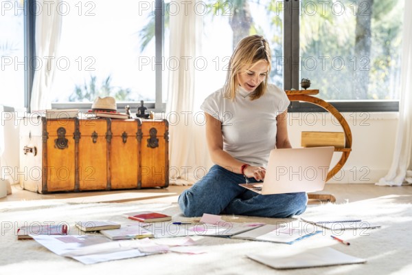 A woman sits on a bright home floor, absorbed in her laptop, with papers scattered around her. Sunlight streams through large windows