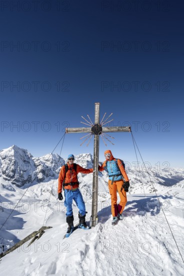 Two ski tourers at the summit cross on the summit of the Madritschspitze, mountain panorama with snow-covered mountain landscape in winter, view of mountain peaks Königsspitze, Monte Zebru and Ortler, Ortler Alps, Vinschgau Valley, Italy