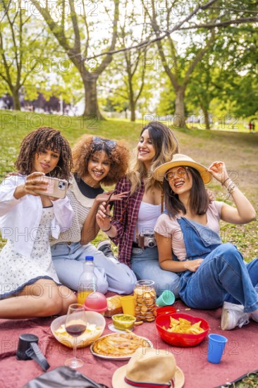 Four cheerful young women enjoying a sunny picnic in the park, happily taking a selfie with their smartphone while surrounded by nature