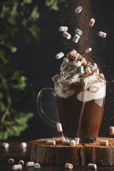 Marshmallows and cocoa powder fall onto a cup of hot chocolate and whipped cream on a wooden board with in a cosy atmosphere