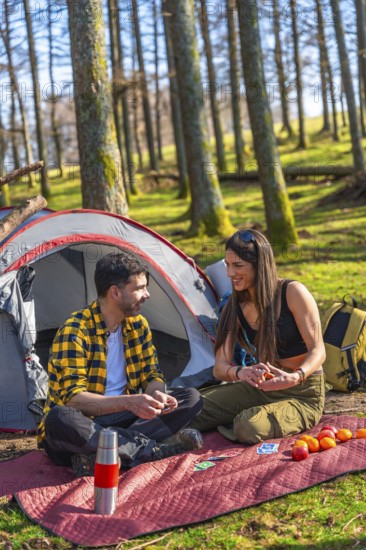Couple playing cards near tent and backpack in forest during camping trip, enjoying sunny day in nature