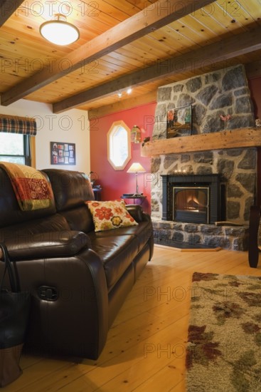 Lit wood burning dark grey natural stone fireplace and brown leather sofa in living room with hardwood floor inside Canadiana style log cabin home, Quebec, Canada