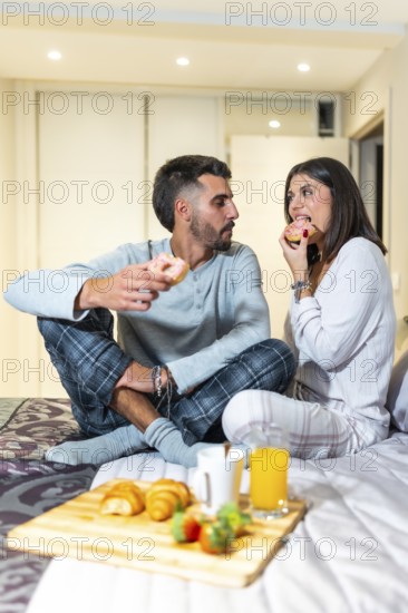 Young couple wearing pajamas sitting in bed eating donuts for breakfast, with croissants, orange juice, strawberries, and coffee on a wooden tray