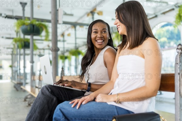 Two young businesswomen are sitting on a bench, working on a laptop and talking, in a light and airy indoor space