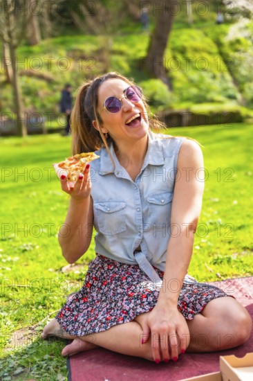Cheerful young woman laughing while eating pizza in a vibrant green park, enjoying a sunny spring day
