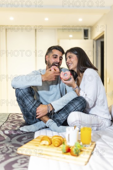 Young couple wearing pajamas, sitting in bed and eating breakfast, including donuts, croissants, orange juice and strawberries