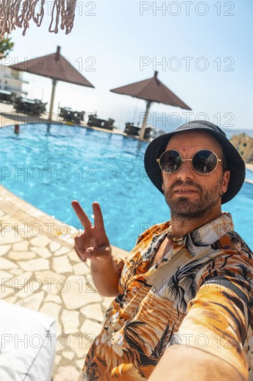 Tourist man in the hotel pool in summer taking a selfie enjoying the vacation, making the victory symbol